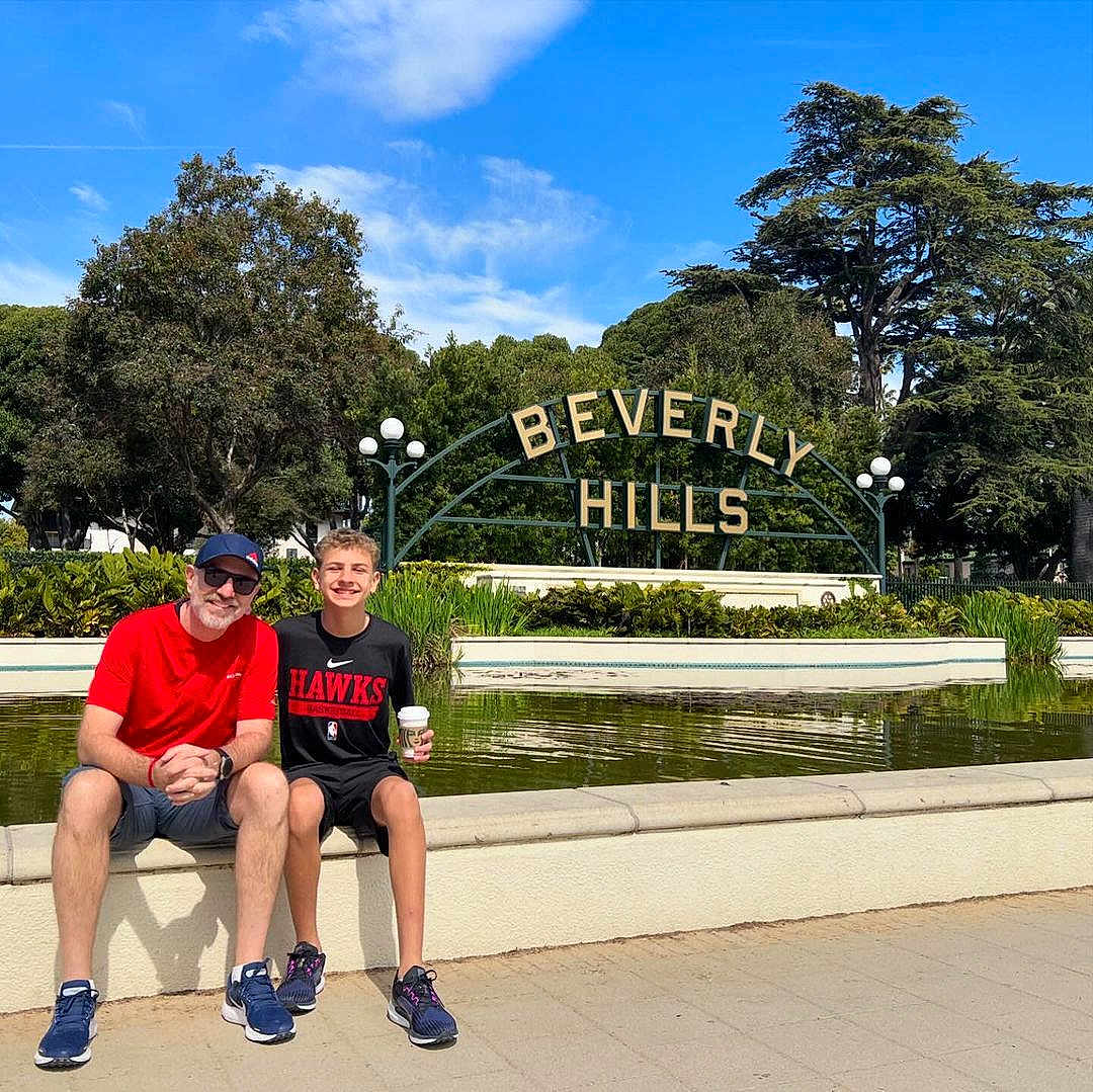 James is registered to the contest to win money with this photo: adult, boy, smiling, sitting, fountain, beverly_hills_sign, outdoor, daytime, trees, greenery, sky, casual_clothing, shorts, sneakers, glasses, cap, coffee_cup, tourist, landmark, urban