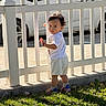 toddler, child, white_picket_fence, grass, sunlight, outdoor, car, curious, shorts, tshirt, socks, shoes, side_view, standing, daytime, shadow, nature, portrait, playful, young_child