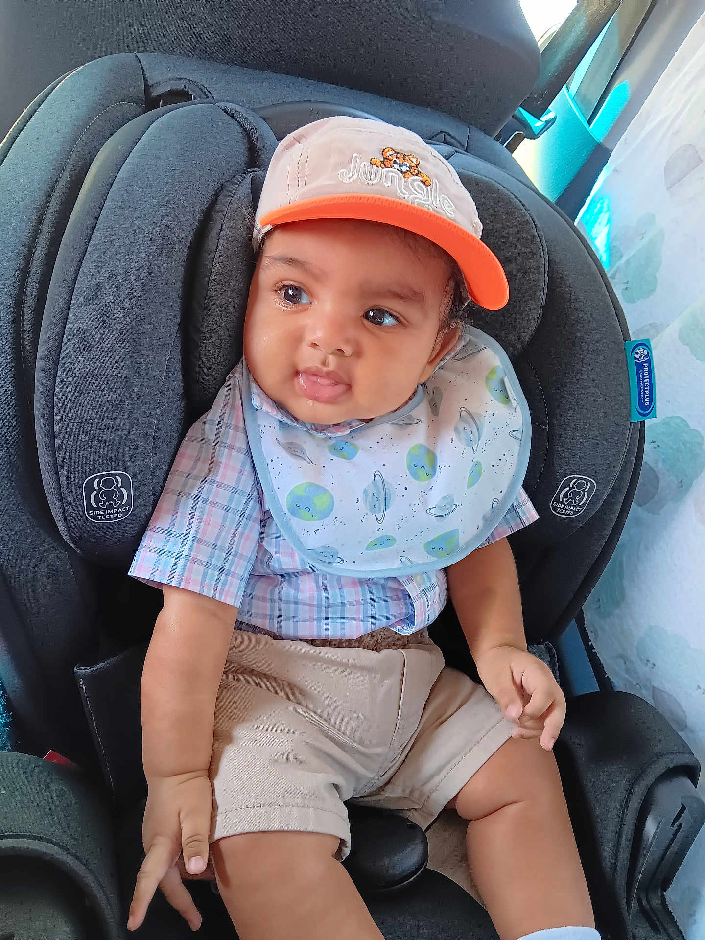 Kayleb is registered to the contest to win money with this photo: baby, child, car_seat, cap, bib, shorts, plaid_shirt, indoor, cute, smiling, person, seatbelt, safety, infant, portrait, clothing, child_seat, happy, baby_face, casual_wear