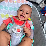 Belen is registered to the contest to win money with this photo: baby, child, smiling, seat, baby_seat, harness, clothing, orange_shirt, indoor, happy, infant, person, cute, comfort, childcare, baby_toy, fabric, pattern, skin, face
