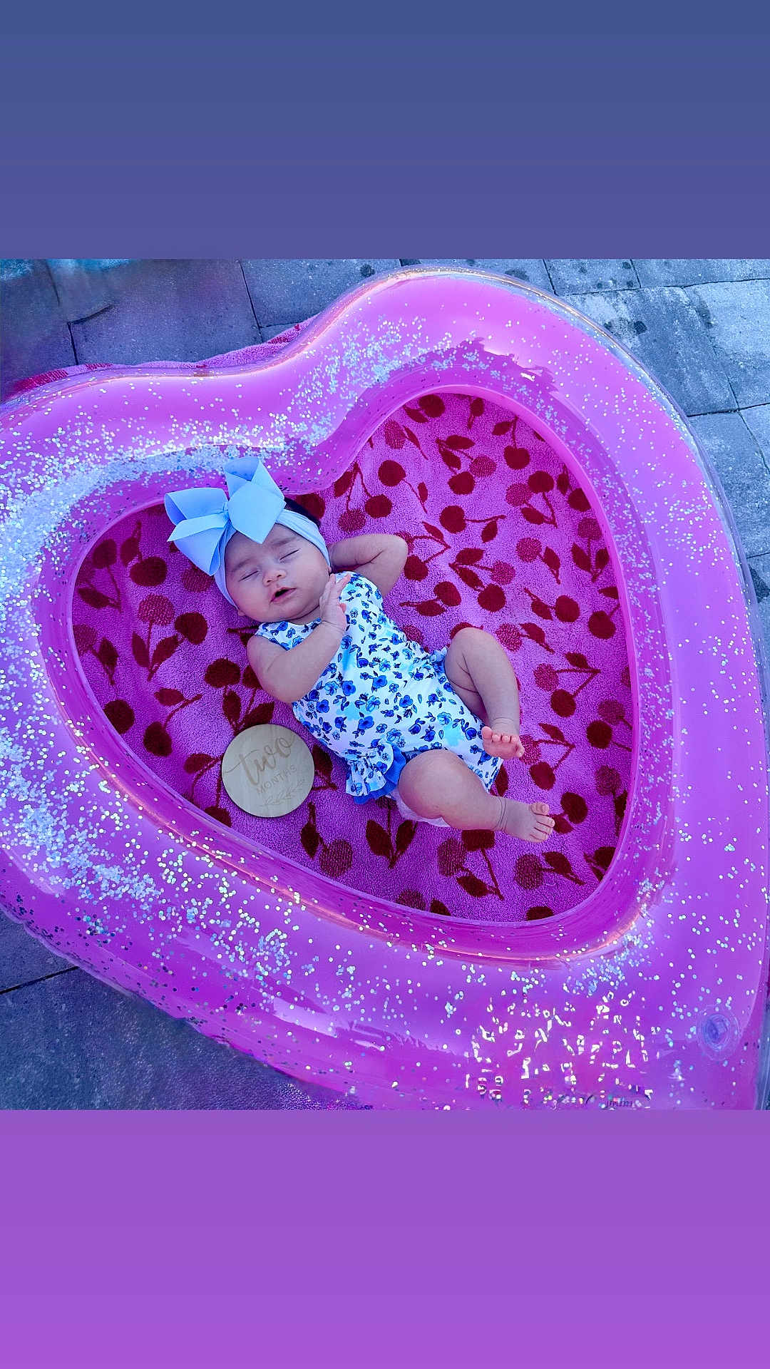 Alanna is registered to the contest to win money with this photo: baby, infant, sleeping, heart_shaped_pool, inflatable, pink, blue_bow, floral_outfit, towel, cherry_pattern, outdoor, pavement, wooden_plaque, age_marker, cute, peaceful, child, portrait, sparkly, relaxing