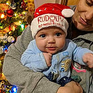 Thomas is registered to the contest to win money with this photo: baby, child, adult, christmas_tree, holiday, festive_hat, rudolph, red_nose, reindeer, smiling, blue_clothing, indoor, warm_lighting, celebration, family, portrait, holding, face, eyes, happy