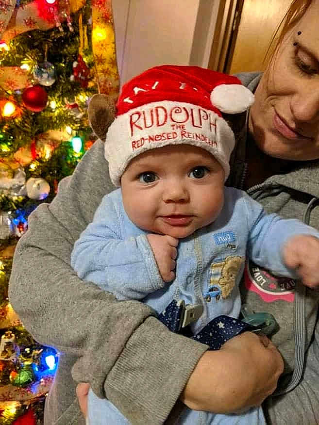 Thomas is registered to the contest to win money with this photo: baby, child, adult, christmas_tree, holiday, festive_hat, rudolph, red_nose, reindeer, smiling, blue_clothing, indoor, warm_lighting, celebration, family, portrait, holding, face, eyes, happy