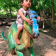 Hanish is registered to the contest to win money with this photo: child, toddler, dinosaur_statue, playground, outdoor, forest, trees, sunlight, sand, barefoot, curious, green, blue, sculpture, nature, play, summer, daylight, young_child, adventure