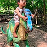 child, toddler, dinosaur_statue, playground, outdoor, forest, trees, sunlight, sand, barefoot, curious, green, blue, sculpture, nature, play, summer, daylight, young_child, adventure