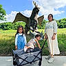 child, woman, dragon_statue, wagon, outdoor, grass, tree, sky, cloud, person, playful, greenery, nature, summer, casual_clothing, sidewalk, smiling, family, fun, sculpture