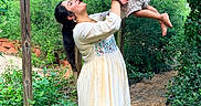 Hanish is registered to the contest to win money with this photo: woman, child, garden, greenery, plants, pathway, wooden_beam, dress, playful, outdoor, nature, happy, smile, holding, barefoot, sunlight, casual_clothing, family, portrait, interaction
