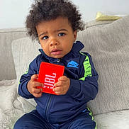 Ishaq a rejoint le concours — aidez-le/la à gagner de superbes lots ! toddler, child, nike, tracksuit, jbl, speaker, couch, indoor, curly_hair, sitting, black_socks, cozy, beige, fabric, young_child, person, clothing, expression, holding, furniture