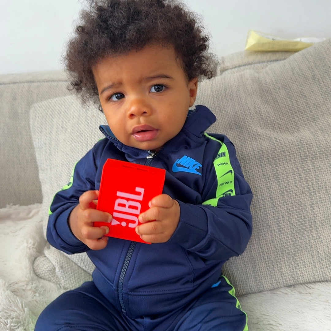 Ishaq a rejoint le concours — aidez-le/la à gagner de superbes lots ! beige, black_socks, child, clothing, couch, cozy, curly_hair, expression, fabric, furniture, holding, indoor, jbl, nike, person, sitting, speaker, toddler, tracksuit, young_child