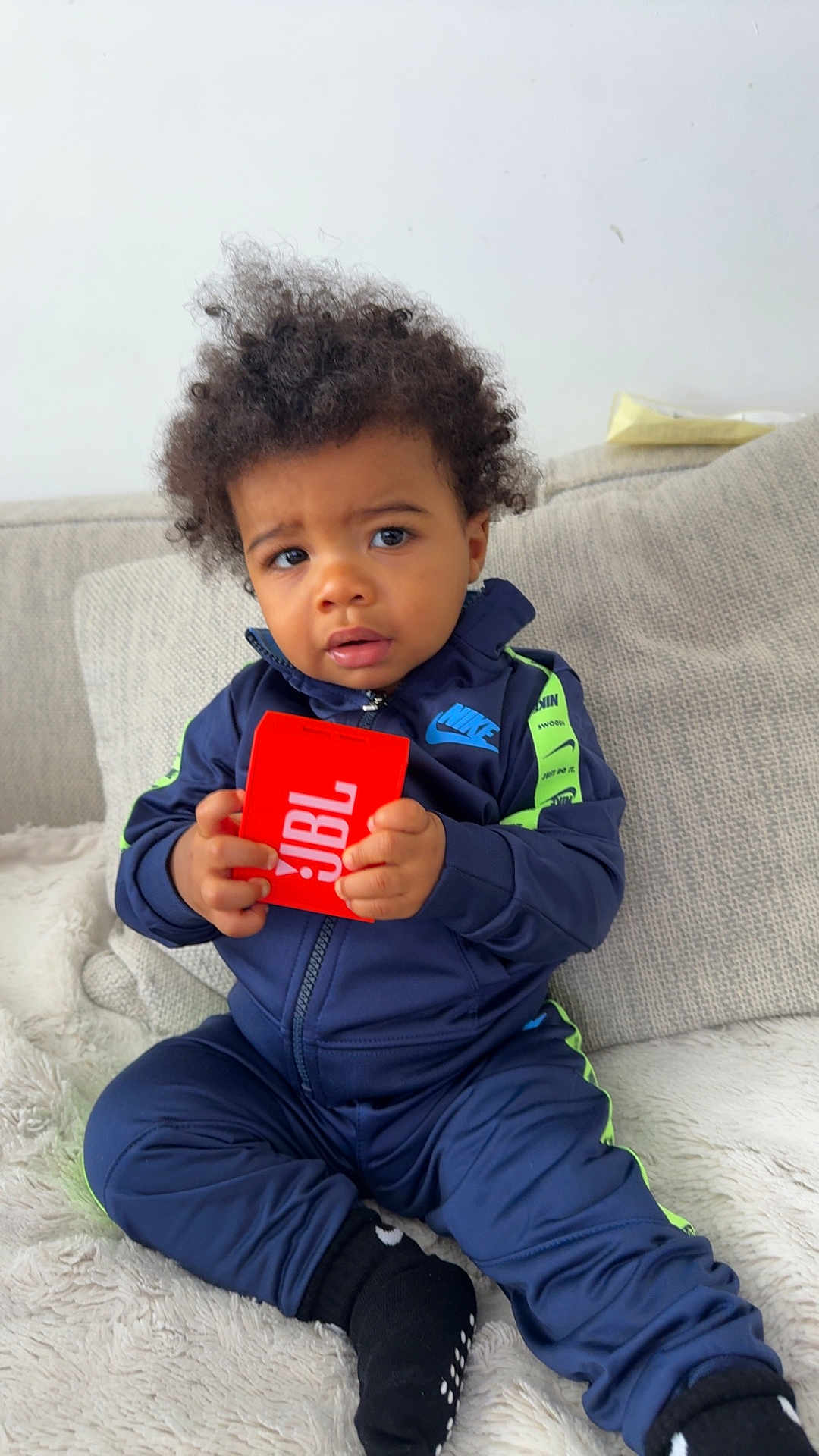 Ishaq a rejoint le concours — aidez-le/la à gagner de superbes lots ! toddler, child, nike, tracksuit, jbl, speaker, couch, indoor, curly_hair, sitting, black_socks, cozy, beige, fabric, young_child, person, clothing, expression, holding, furniture
