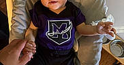 Kenzo is registered to the contest to win money with this photo: baby, infant, beanie, purple_shirt, logo, baby_seat, hand_holding, adult_hand, wooden_floor, pet_bowls, cushion, indoor, person, smile, cute, child, clothing, portrait, home, comfortable