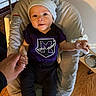baby, infant, beanie, purple_shirt, logo, baby_seat, hand_holding, adult_hand, wooden_floor, pet_bowls, cushion, indoor, person, smile, cute, child, clothing, portrait, home, comfortable