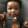 baby, child, face, curly_hair, onesie, pumpkin, ghost, halloween, cute, portrait, infant, smiling, hand, clothing, lying_down, indoors, soft_surface, black_hair, closeup, person
