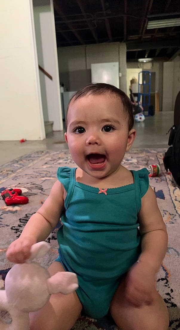 Luna is registered to the contest to win money with this photo: baby, baby_toddler_clothing, chair, cheek, child, event, facial_expression, finger, flooring, fun, happy, human, iris, mouth, nail, person, sitting, skin, standing, thumb