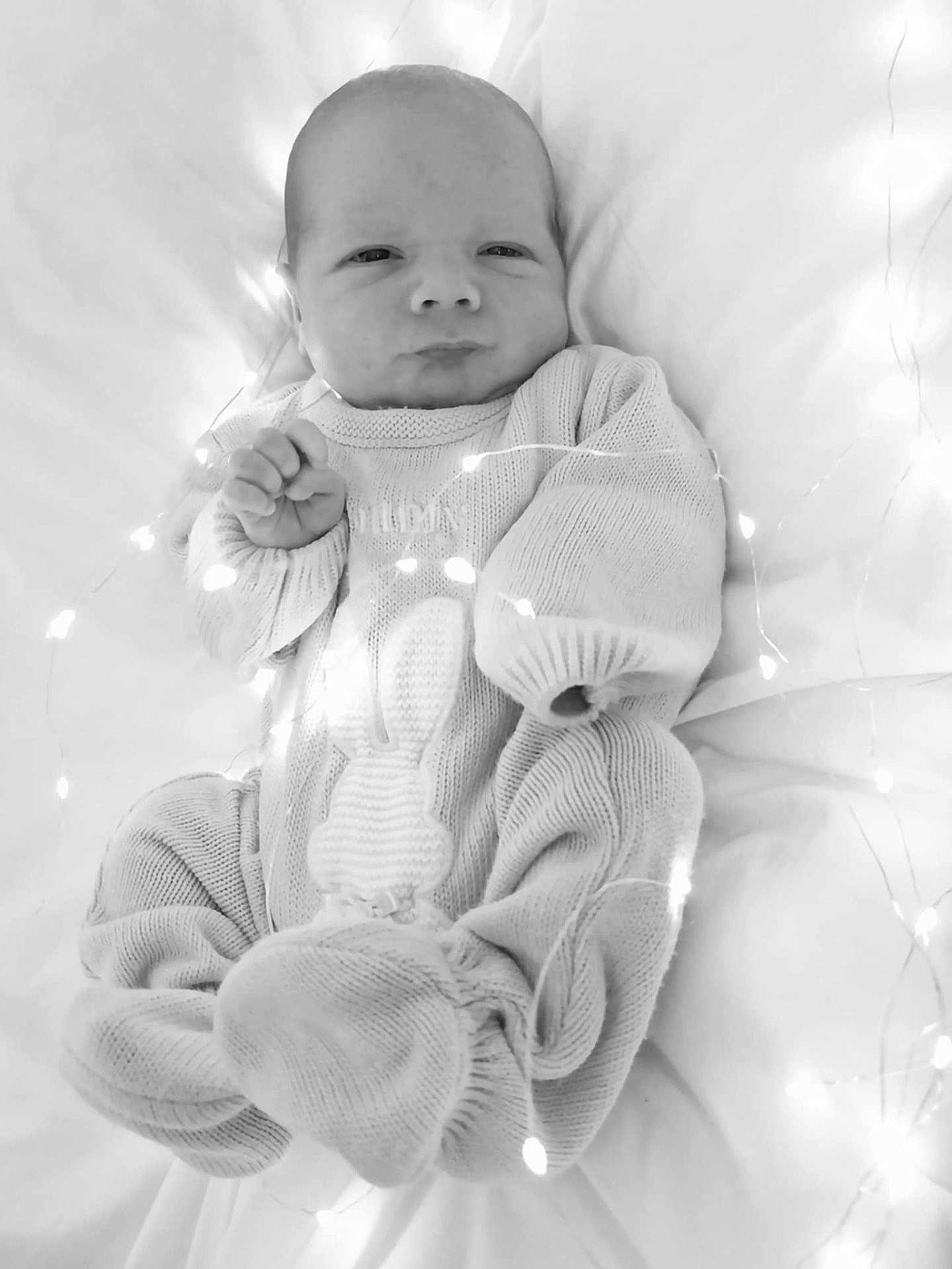 Tommy is registered to the contest to win money with this photo: baby, infant, knitwear, blanket, fairy_lights, cozy, cute, portrait, newborn, soft, white, sleepwear, child, face, expression, hands, lying_down, indoors, warm, light