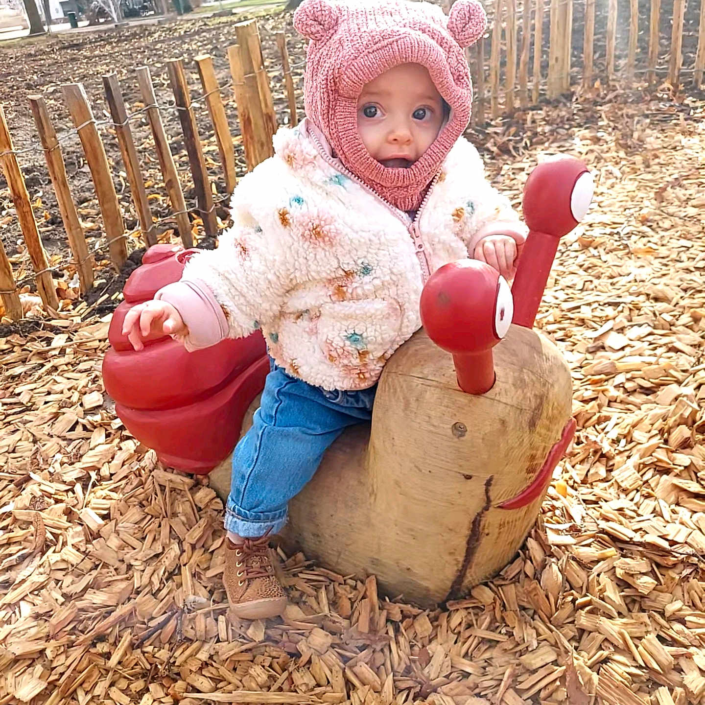Capucine a rejoint le concours — aidez-le/la à gagner de superbes lots ! bear_ears, blue_jeans, brown_boots, child, curious, cute, daylight, fall, fence, fluffy_jacket, leafless_trees, outdoor, park, pink_hat, playground, sitting, toddler, toy, wood_chips, wooden_snail