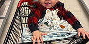 Bennett is registered to the contest to win money with this photo: baby, shopping_cart, blanket, pajamas, child, smiling, indoor, retail_store, aisle, floor, person, jeans, boots, hand, face, head, clothing, metal, furniture, happy