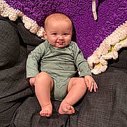 Bennett is registered to the contest to win money with this photo: baby, child, infant, smiling, tongue_out, cushion, blanket, purple_blanket, cream_blanket, green_onesie, dinosaur_print, sitting, indoors, cute, happy, skin, face, foot, hand, cozy