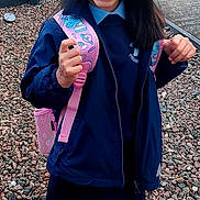 Olivia joined the competition — help win amazing prizes! child, girl, smiling, yellow_bows, school_uniform, backpack, outdoor, rocks, fence, grass, black_shoes, white_socks, happy, portrait, standing, person, school, primary_school, daylight, casual