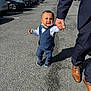 child, toddler, boy, suit, bow_tie, walking, hand_holding, adult, parking_lot, cars, outdoor, pavement, sunlight, shadow, formal_clothing, smile, person, footwear, nature, greenery