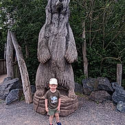 Maël participe au concours pour gagner de l'argent avec cette photo : boy, casual_clothing, child, daylight, forest, gravel, hat, nature, outdoor, park, playful, rocks, sculpture, shorts, smile, sneakers, standing, statue, trees, wooden_bear