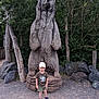 boy, casual_clothing, child, daylight, forest, gravel, hat, nature, outdoor, park, playful, rocks, sculpture, shorts, smile, sneakers, standing, statue, trees, wooden_bear