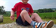 Maël a rejoint le concours — aidez-le/la à gagner de superbes lots ! cap, casual, child, clouds, daylight, field, grass, greenery, happy, hay_bale, nature, outdoor, portrait, red_shirt, rural, shorts, sitting, sky, smiling, sneakers