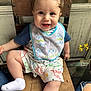 baby, child, smiling, bib, dinosaur_print, wooden_chair, drool, cute, happy, sitting, indoor, clothing, shorts, sock, person, portrait, hair, expression, yellow_flower, rustic_background