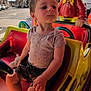 child, toddler, ride, kiddie_ride, car, toy, amusement, outdoor, street, building, daylight, person, clothing, shorts, shirt, footwear, plastic, colorful, playful, urban