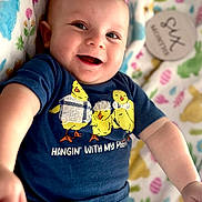 Levi is registered to the contest to win money with this photo: baby, smiling, infant, blue_shirt, yellow_chicks, text_on_clothing, colorful_blanket, patterned_blanket, happy, cute, portrait, indoor, child, person, closeup, laying_down, soft_lighting, casual_clothing, playful, young_child