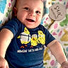 baby, smiling, infant, blue_shirt, yellow_chicks, text_on_clothing, colorful_blanket, patterned_blanket, happy, cute, portrait, indoor, child, person, closeup, laying_down, soft_lighting, casual_clothing, playful, young_child