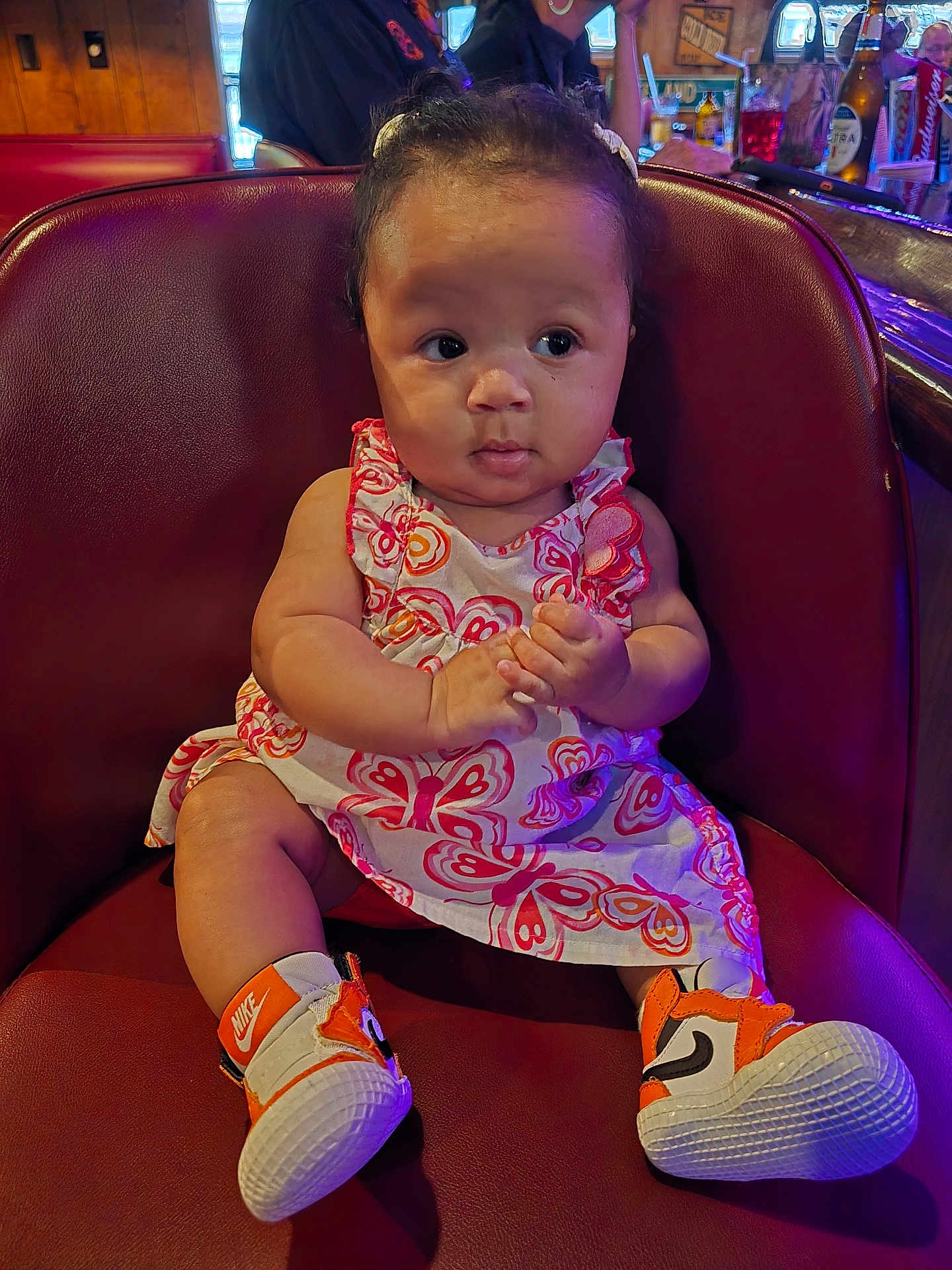 Clover is registered to the contest to win money with this photo: baby, child, sitting, dress, butterfly_pattern, sneakers, nike, orange, curly_hair, leather_chair, indoor, restaurant, bar, bottle, glass, person, cute, hands, expression, casual