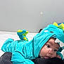 baby, child, dinosaur_costume, onesie, blue, green, spikes, cute, infant, black_cushion, tongue_out, lying_down, indoor, soft_fabric, costume, face, hand, expression, wall, bed