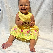 Normani joined the competition — help win amazing prizes! baby, child, smile, yellow_dress, flower_embroidery, wooden_plaque, 10_months, happy, sitting, barefoot, earrings, blanket, indoor, portrait, cute, infant, person, celebration, teeth, skin