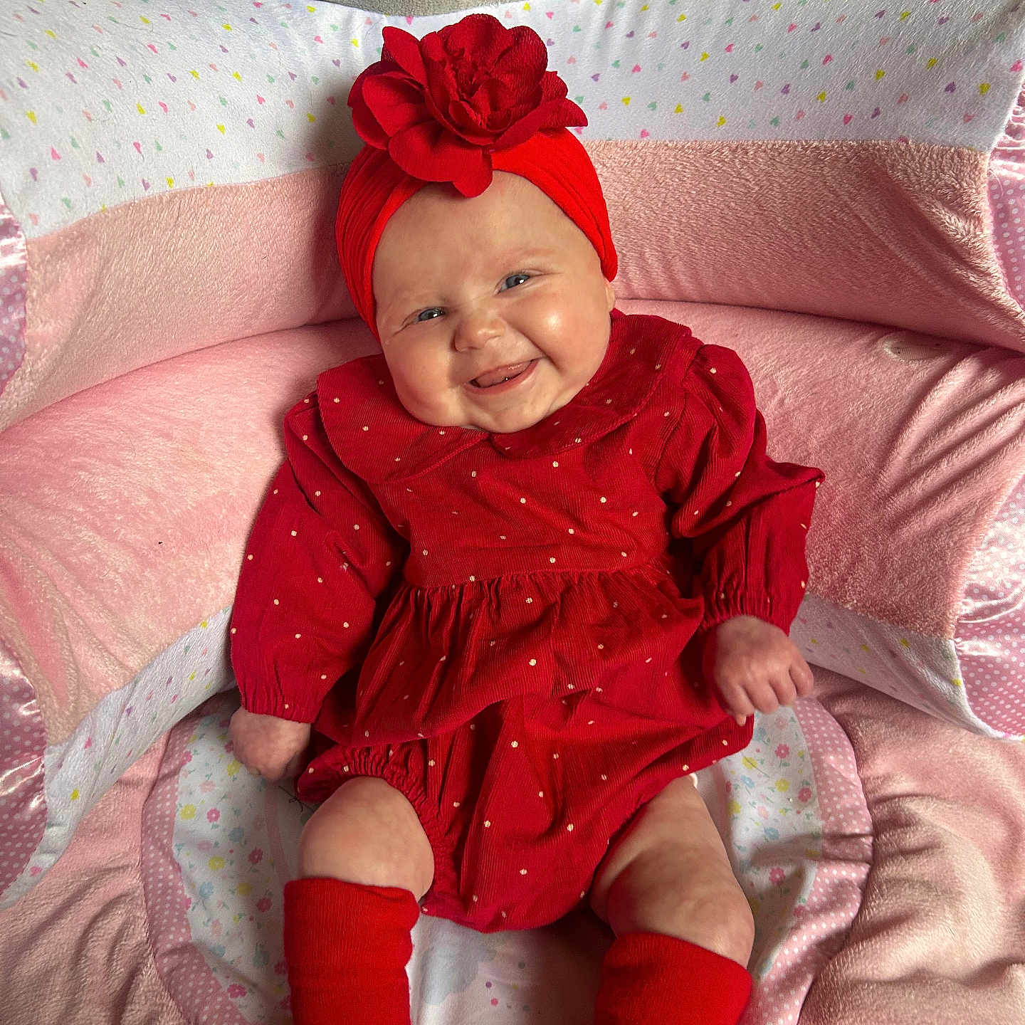 Bella is registered to the contest to win money with this photo: baby, child, comfortable, cozy, cushion, cute, face, flower, happy, headband, indoors, infant, pink, plush, portrait, red_clothing, sitting, smile, sock, soft_toys