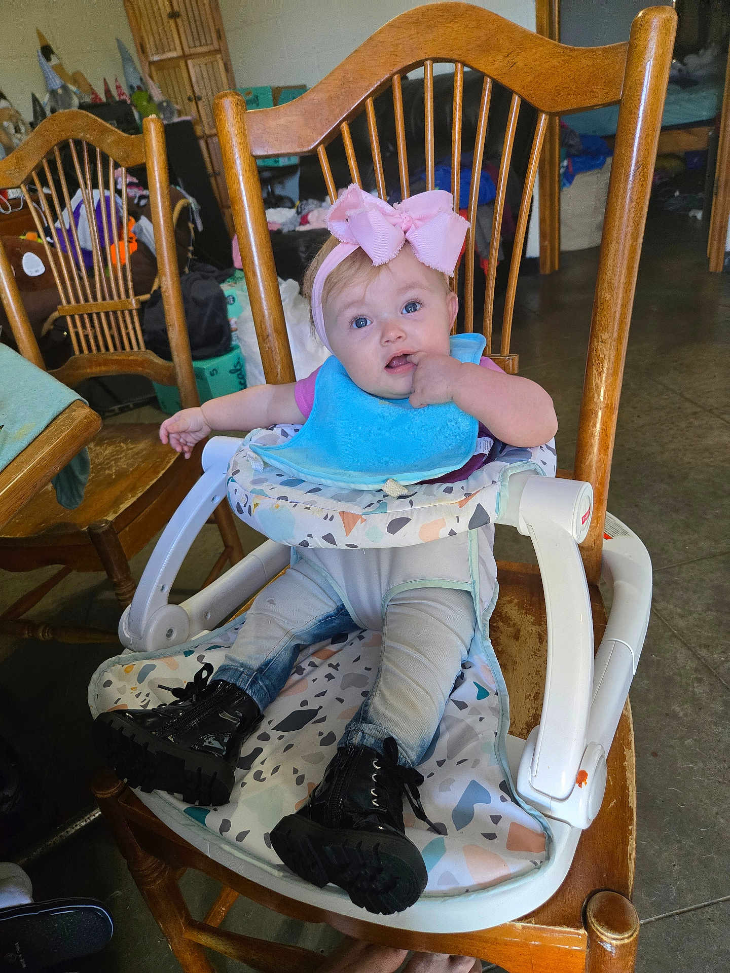 Zailee is registered to the contest to win money with this photo: baby, child, highchair, pink_bow, bib, black_boots, jeans, wooden_chair, indoor, portrait, face, blue_eyes, hand_in_mouth, feeding_seat, clothing, floor, household, smile, cute, messy_background