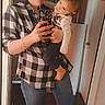 woman, toddler, child, mirror, selfie, indoor, casual_clothing, plaid_shirt, jeans, barefoot, phone, smiling, holding_child, curly_hair, wooden_floor, door, home, portrait, happy, family