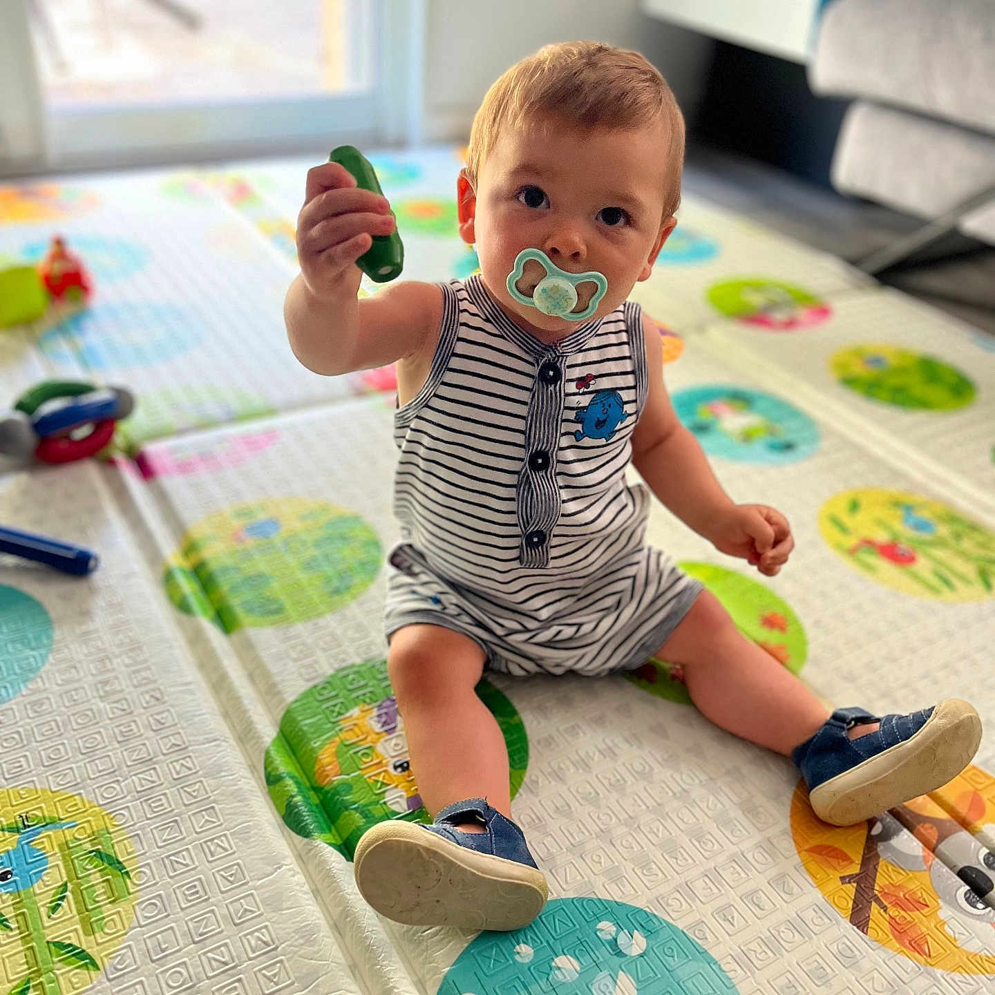 Louis participe au concours pour gagner de l'argent avec cette photo : baby, carpet, child, colorful, curious, cute, floor, footwear, home, indoor, living_room, pacifier, person, playmat, sitting, striped_clothing, toddler, toy, window, young_child