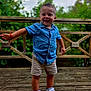 boy, bracelet, casual_clothing, child, daytime, denim_shirt, feet, greenery, happy, natural_light, outdoor, playful, portrait, short_hair, shorts, smiling, sneakers, standing, toddler, wooden_deck