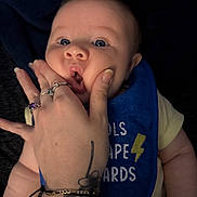 Ronin is registered to the contest to win money with this photo: adult_hand, baby, bib, blue_eyes, cheeks, closeup, clothing, cute, dark_background, expression, face, hand, infant, jewelry, person, playful, rings, skin, tattoo, yellow_shirt