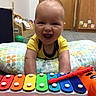 baby, child, infant, smiling, happy, laughing, face, portrait, sitting, toy, xylophone, musical_instrument, colorful, playroom, indoor, cushion, playmat, yellow_shirt, wooden_door, close_up