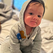 Kyan is registered to the contest to win money with this photo: baby, bed, blurred_background, casual_clothing, child, cozy, curious, cute, face, grey_clothing, hoodie, indoor, person, portrait, sitting, smile, soft_light, toddler, young_child, youth
