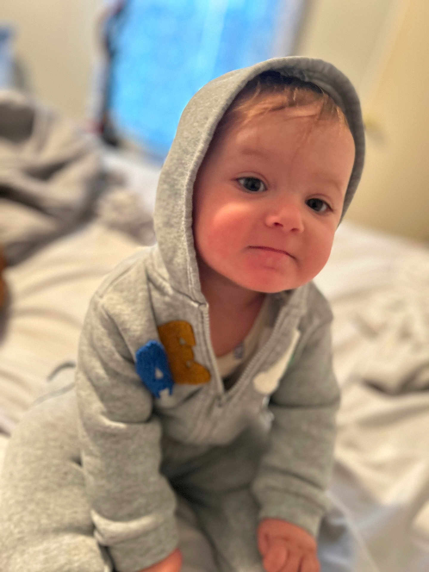 Kyan is registered to the contest to win money with this photo: baby, bed, blurred_background, casual_clothing, child, cozy, curious, cute, face, grey_clothing, hoodie, indoor, person, portrait, sitting, smile, soft_light, toddler, young_child, youth