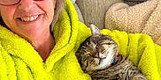 Grayson joined the competition — help win amazing prizes! woman, cat, glasses, hoodie, grinch, couch, blanket, smiling, happy, cozy, indoor, tabby_cat, pet, relaxed, sleepy, furniture, portrait, casual, comfort, friendship