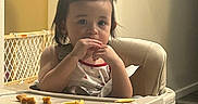 Alexus is registered to the contest to win money with this photo: toddler, child, high_chair, food, snack, indoor, hair, pony_tail, face, person, baby_gate, wall, floor, table, seat, clothing, cute, eating, light, furniture