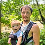 Kohen is registered to the contest to win money with this photo: baby, blurred_background, carrier, casual_clothing, child, daylight, forest, greenery, happy, man, nature, outdoor, parent_and_child, person, portrait, short_sleeves, smile, summer, tattoo, trees