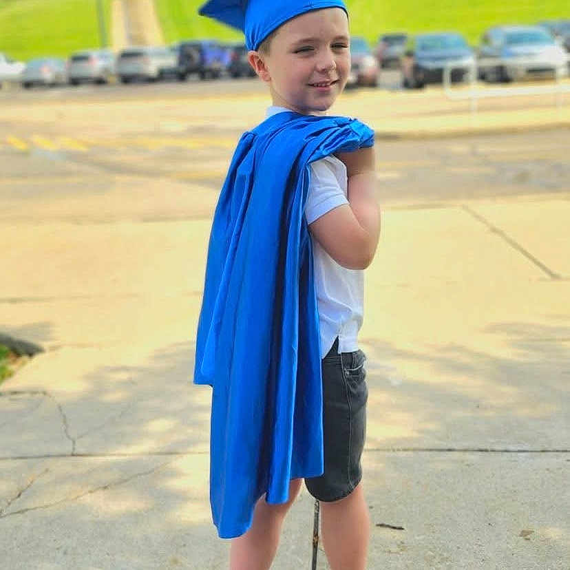 Liam is registered to the contest to win money with this photo: achievement, blue, building, cap, cars, celebration, child, daytime, gown, graduation, grass, outdoor, person, portrait, school, shoes, shorts, sidewalk, smile, socks