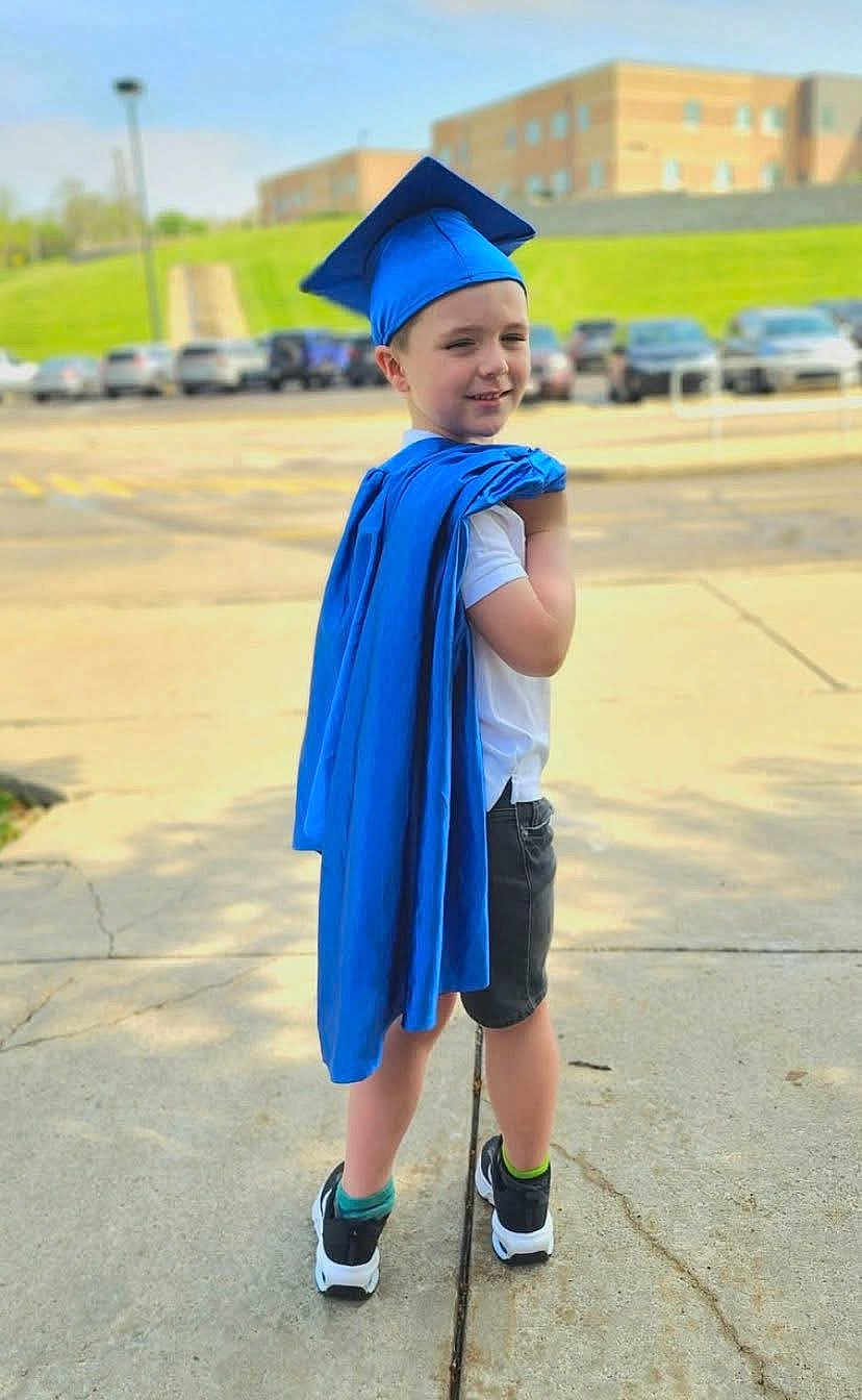 Liam is registered to the contest to win money with this photo: child, graduation, cap, gown, blue, sidewalk, outdoor, smile, shorts, shoes, socks, school, building, cars, daytime, grass, celebration, portrait, person, achievement