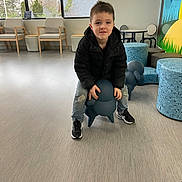 Liam is registered to the contest to win money with this photo: child, boy, black_jacket, ripped_jeans, sneakers, indoor, waiting_room, stool, blue, dog_shaped, furniture, chairs, table, mural, floor, window, daylight, playful, smile, casual