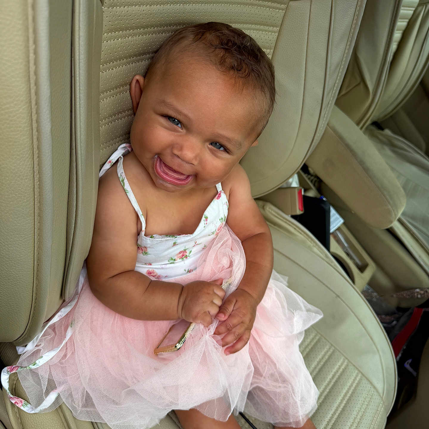 Azalea is registered to the contest to win money with this photo: baby, child, smiling, tutu, pink_dress, car_seat, beige_leather, happy, cute, portrait, sitting, person, infant, curly_hair, barefoot, holding_object, closeup, indoor, seatbelt, car_interior